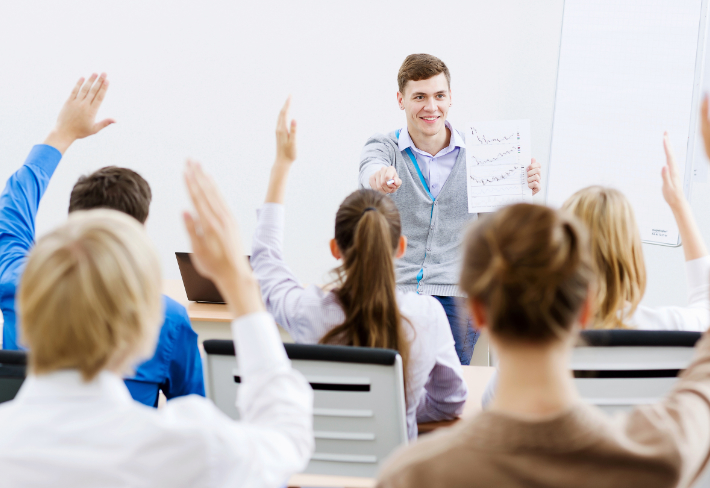 Na pierwszym planie nauczyciel, a w tle uczniowie podnoszący rękę.