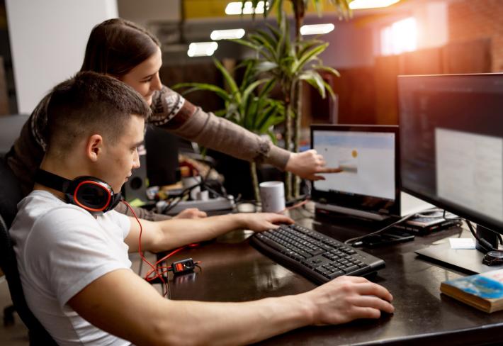 Na zdjęciu dwoje młodych (chłopak i dziewczyna) ludzi przy komputerze. Dziewczyna pokazuje chłopakowi coś na monitorze.